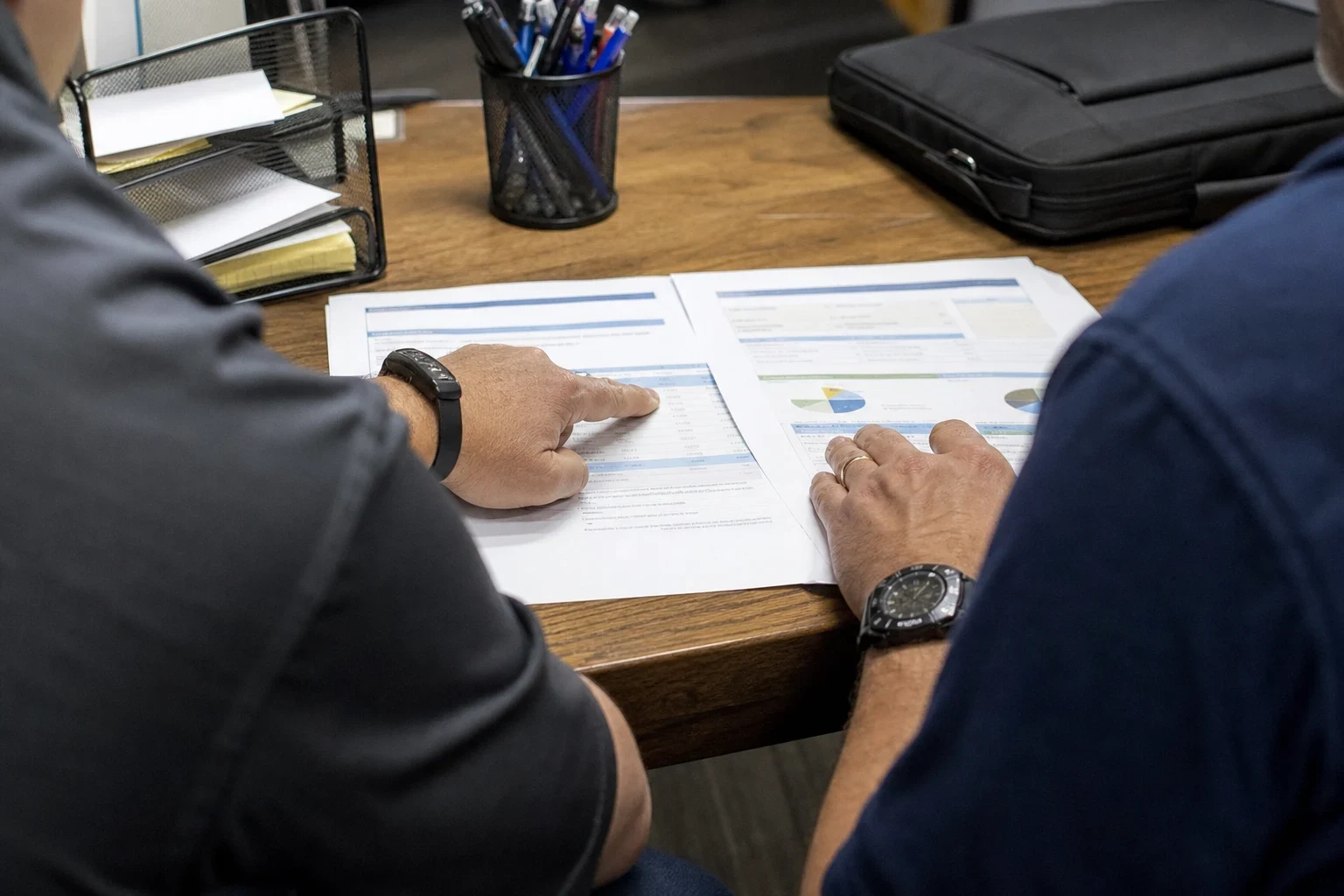 Over-the-shoulder view of two men at a wooden office desk comparing two printed documents with tables and pie charts, beside a pen cup, paper tray and laptop bag